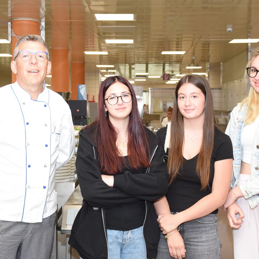 Vier Personen stehen in einer Küche. Ein Mann in einem Kochuniform, zwei junge Frauen und eine weitere Frau mit langen blonden Haaren. Sie lächeln und scheinen für ein Foto zu posieren.