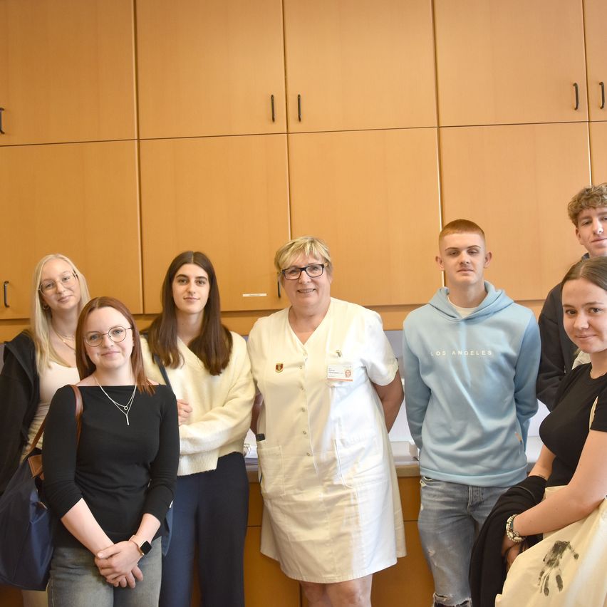 Eine Gruppe von Menschen, möglicherweise Studenten, steht vor einem Holzschrank. Sie alle lächeln. Die Frau in der Mitte trägt einen weißen Kittel.