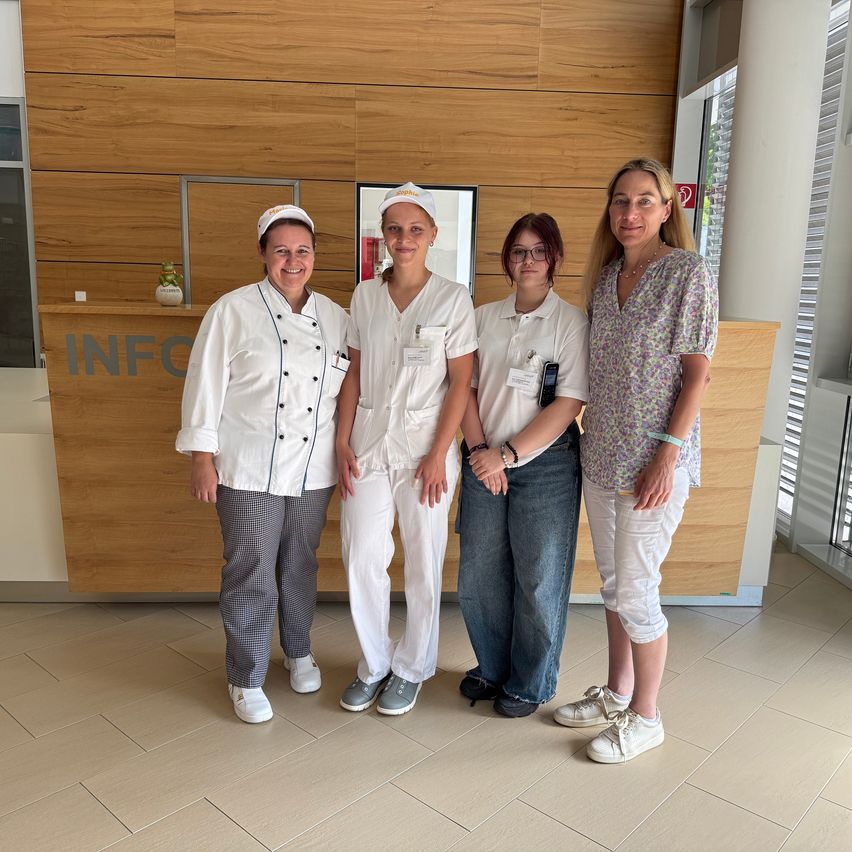 Fünf Frauen stehen zusammen in einem gut beleuchteten Raum, jede unterschiedlich gekleidet. Sie stehen vor einem hölzernen Empfangstresen mit der Aufschrift INFO.