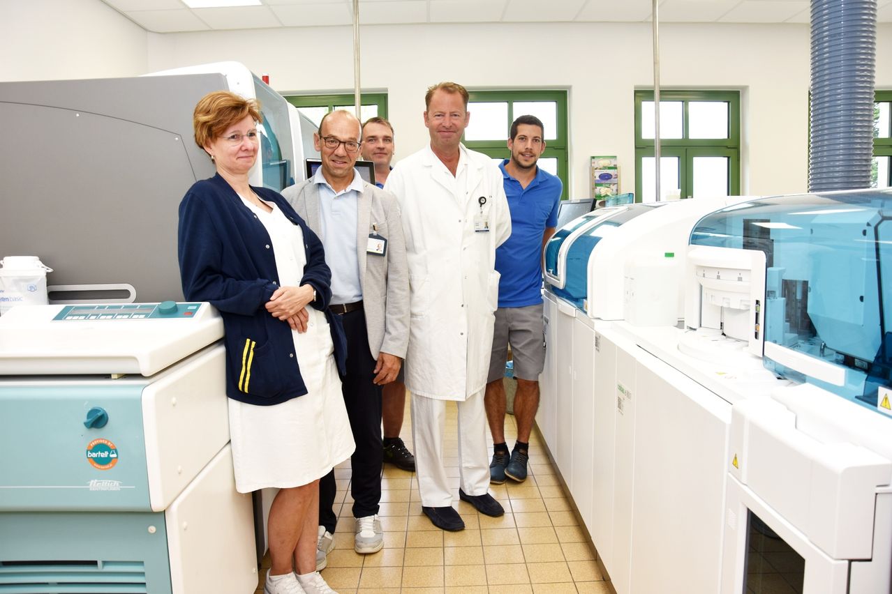 Fünf Personen stehen in einem Raum, vermutlich einem Labor. Der Mann in Weiß trägt einen Laborkittel und eine Namensplakette. Die Frau links trägt ein weißes Kleid. Die anderen Personen sind lässig gekleidet. Hinter ihnen stehen weiße Maschinen.