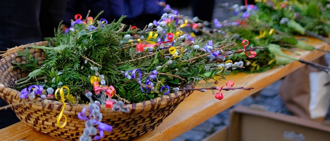 Ein hölzerner Tisch mit einem Korb voller grüner Blumen und bunten Schleifen. Hinter dem Tisch sind verschwommene Menschen in Winterkleidung zu sehen.