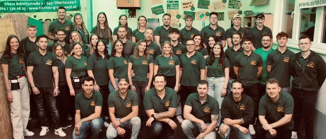 Eine Gruppe von Menschen in grünen Polo-Shirts posiert für ein Foto in einem Raum mit einem Banner, das 'Wald' auf der Wand steht.