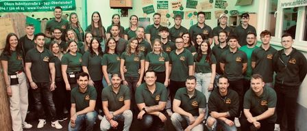 Eine Gruppe von Menschen in grünen Polo-Shirts posiert für ein Foto in einem Raum mit einem Banner, das 'Wald' auf der Wand steht.