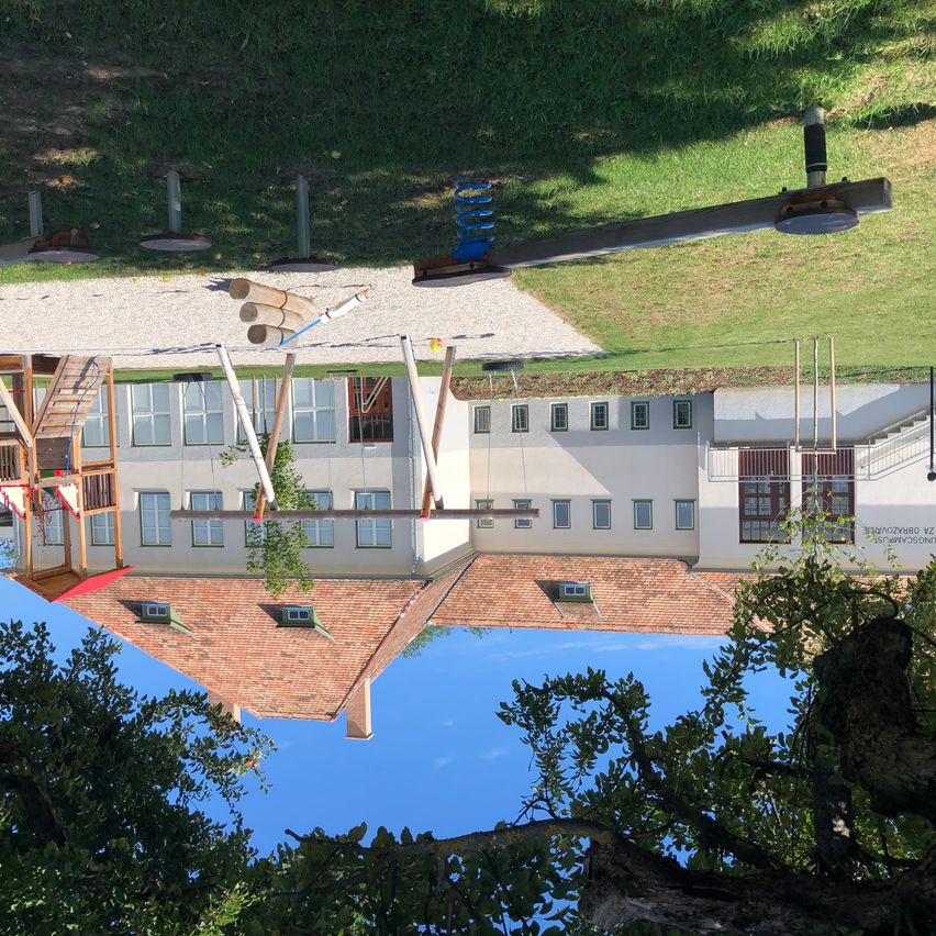Ein Gebäude mit vielen Fenstern und einem roten Dach. Ein Spielplatz mit Rutschen und Leitern davor. Bäume und ein blauer Himmel im Hintergrund.