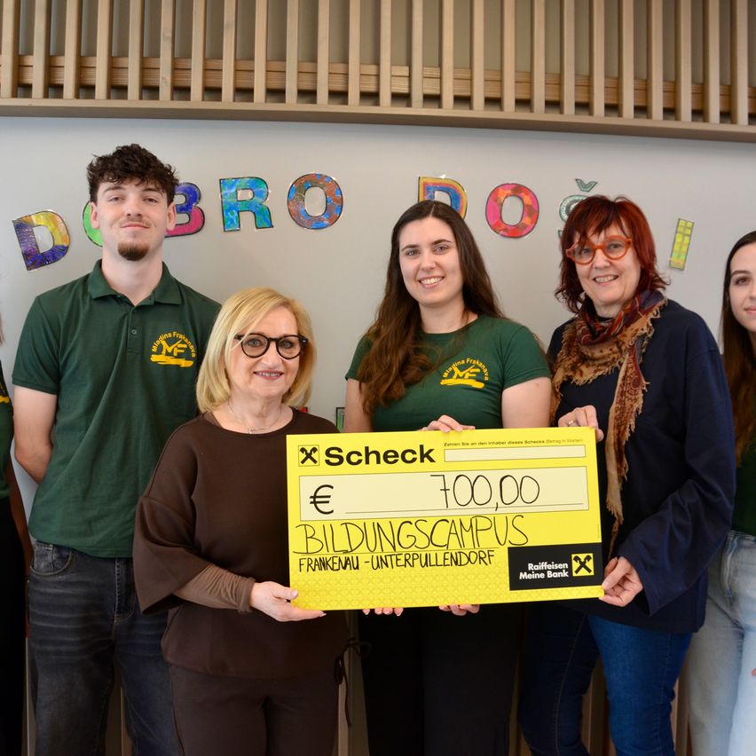 Eine Frau, die einen großen Scheck hält, steht mit vier anderen vor einer Wand mit bunten Buchstaben. Der Scheck ist für 100.000 Euro von Bildungscampus Frankenau-Unterpullendorf.