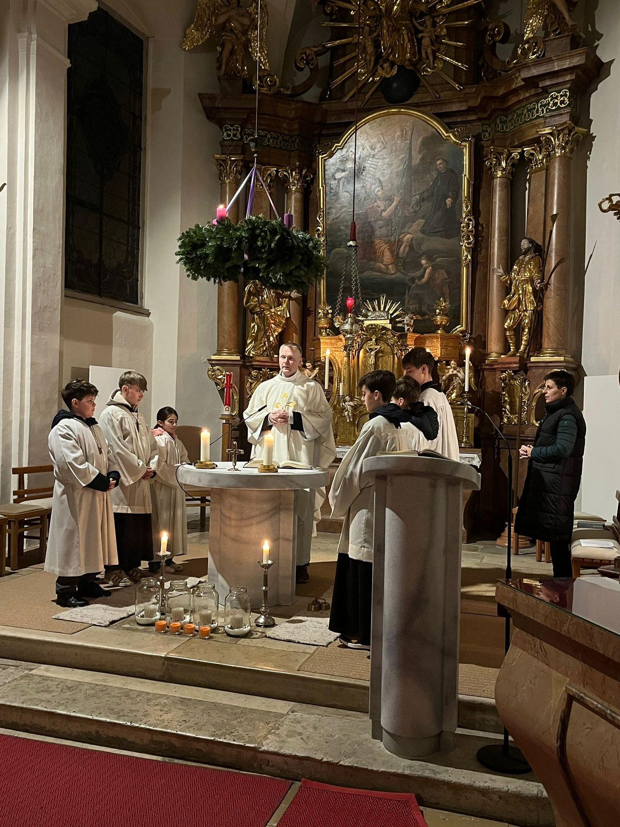 Ein Priester steht vor einem Altar mit Kerzen, umgeben von Ministranten und einer Frau. Hinter ihm schmücken eine goldene Statue und ein Gemälde den Altar.