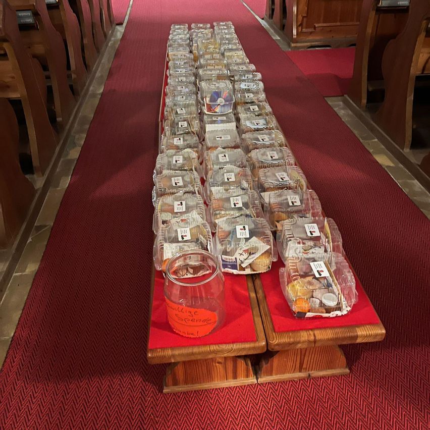 Gang eines Kirchengebäudes mit Reihen von durchsichtigen Plastikbehältern mit Lebensmitteln, einschließlich eines Glases mit der Aufschrift 'Sainte Pierre 1996', auf einem roten Teppich.