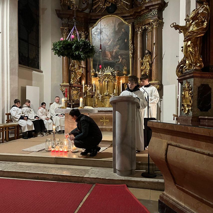 Eine feierliche religiöse Zeremonie findet in einer Kirche statt. Ein Priester zündet Kerzen am Altar an. Eine Gruppe von Menschen sitzt und beobachtet.