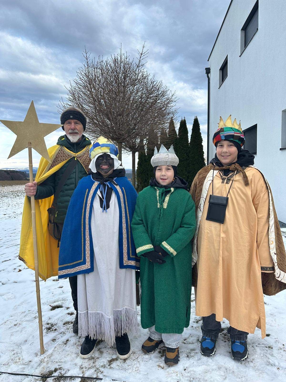 Vier Menschen in Kostümen stehen im Schnee vor einem Gebäude. Der Mann links hält einen Stern, und die anderen tragen Kronen. Der Boden ist mit Schnee bedeckt.