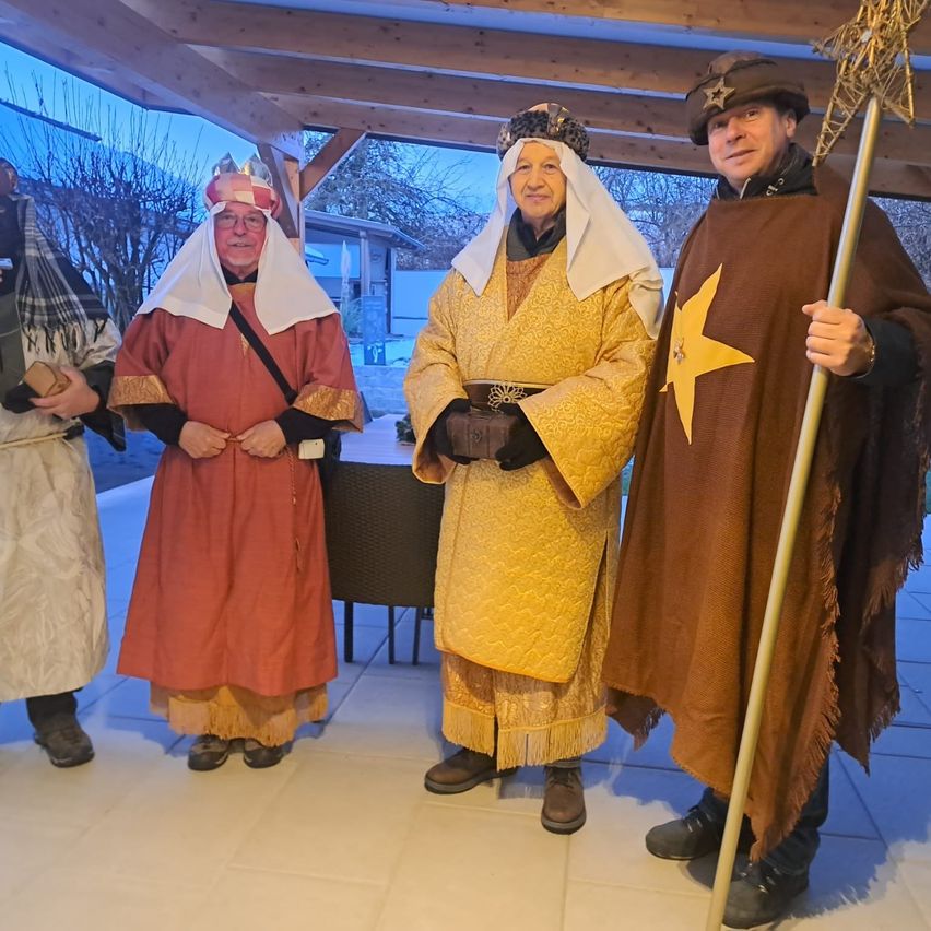 Vier Menschen in Kostümen stehen in einem Raum zusammen. Einer hält einen Stab mit einem Stern, ein anderer trägt eine Krone, und zwei weitere haben Kopftücher. Ein Tisch und Stühle sind im Hintergrund.