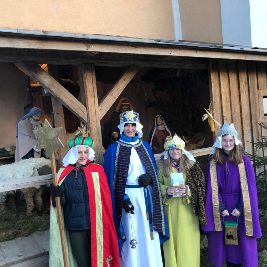 Fünf Personen in aufwendigen Kostümen stehen vor einer Krippe. Einer hält einen Stab und einen Stern, ein anderer trägt eine Krone und einen blauen Umhang, zwei halten Geschenke und die fünfte ist mit einem lila Umhang geschmückt.