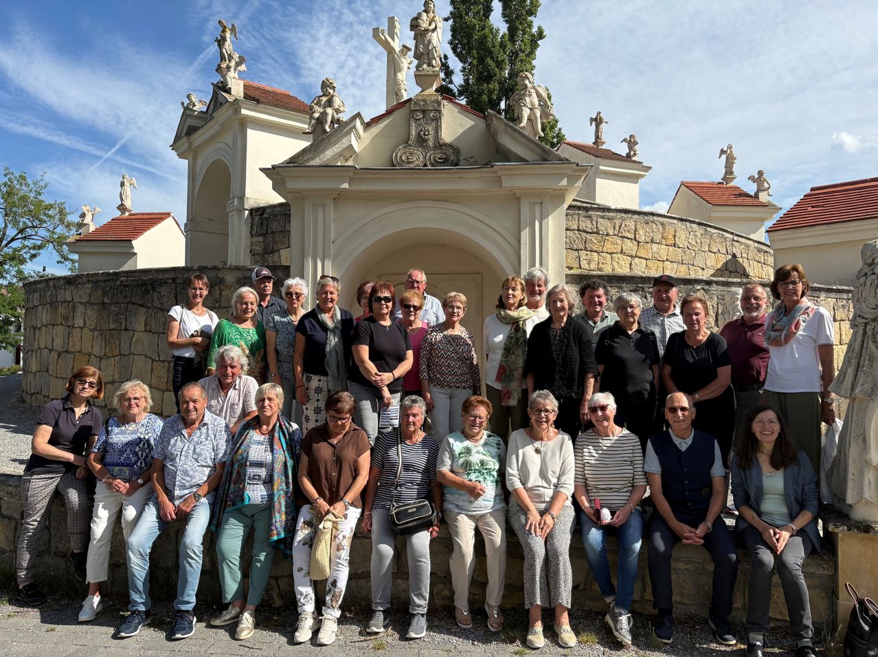 Eine Gruppe von Menschen posiert für ein Foto vor einer historischen Stätte mit Skulpturen auf der Spitze, unter einem blauen Himmel.