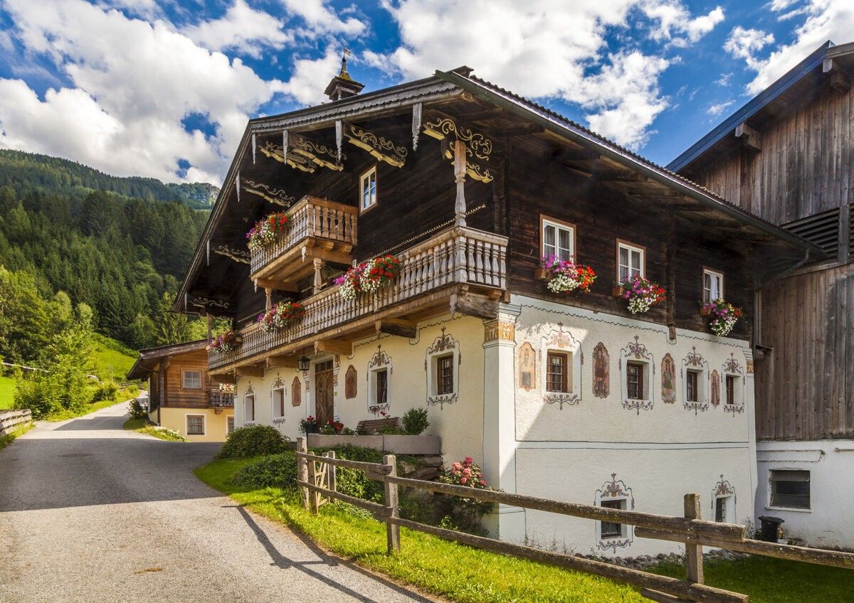 A traditional wooden house with decorative balconies and flower boxes, nestled in a mountainous landscape with a wooden fence in the foreground.