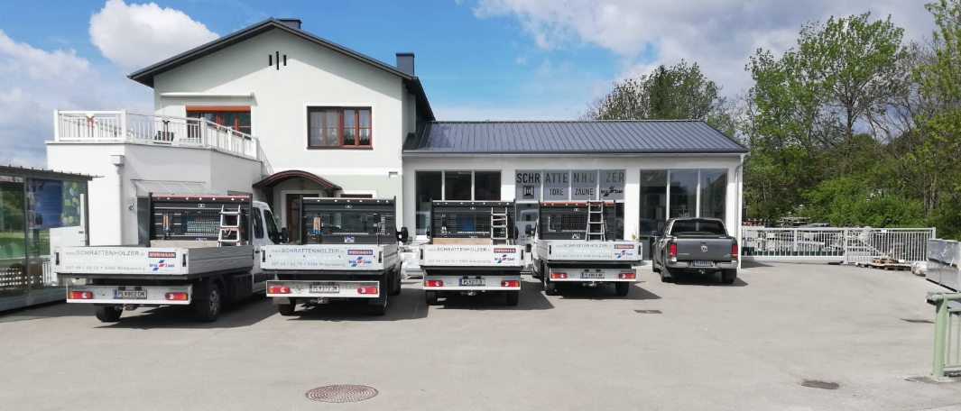 Mehrere Lastwagen sind vor einem weißen Gebäude mit Glasfenstern geparkt. Das Gebäude hat ein Schild mit der Aufschrift 'Schrattenholzer'. Es gibt auch einige Bäume in der Umgebung.