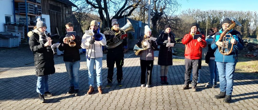 Eine Gruppe von Menschen steht auf einem gepflasterten Weg und hält Musikinstrumente in den Händen. Einige von ihnen spielen Trompeten, während andere Posaunen spielen. Sie führen wahrscheinlich eine Aufführung bei einer Veranstaltung im Freien durch.