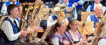 Eine Band spielt auf der Bühne mit verschiedenen Musikinstrumenten. Der Mann in der Mitte spielt Tuba. Die Frau rechts spielt Klarinette. Das Publikum schaut ihnen zu.