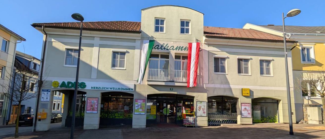 Ein Gebäude mit rotem Dach hat ein Schild mit der Aufschrift 'Alhamn'. Fahnen sind am Gebäude gehisst. Unter dem Schild befindet sich ein Geschäft mit einer Glastür und einer Glaswand. Draußen steht ein Einkaufswagen.