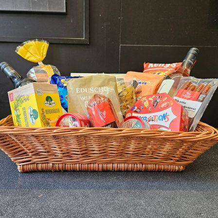 Ein Korb aus Weide auf einem Tresen enthält eine Mischung aus Lebensmitteln, darunter Weinflaschen, eine Schachtel Bergkrauter, eine rote Schachtel, eine Tüte Nüsse und andere Snacks.