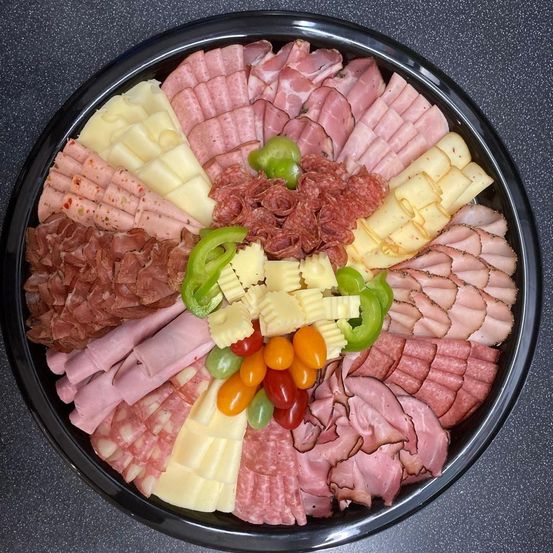 Ein schwarzer Teller enthält eine Auswahl an Fleisch und Käse, mit Scheiben von Salami, Schinken und Pepperoni, zusammen mit Käsestücken und bunten Gemüsen.