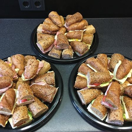 Drei schwarze Teller zeigen Sandwiches mit Sesam, Käse, Tomaten und Schinken. Die Sandwiches sind in Hälften geschnitten und ordentlich angeordnet. Der Hintergrund ist eine dunkle, möglicherweise hölzerne Oberfläche.