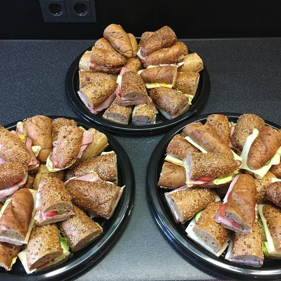 Drei schwarze Teller zeigen Sandwiches mit Sesam, Käse, Tomaten und Schinken. Die Sandwiches sind in Hälften geschnitten und ordentlich angeordnet. Der Hintergrund ist eine dunkle, möglicherweise hölzerne Oberfläche.