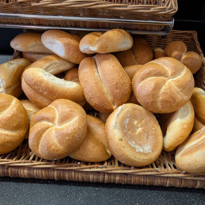 A basket filled with various types of bread, including loaves with twisted tops and others with smooth crusts, is stacked on another basket in a bakery setting.