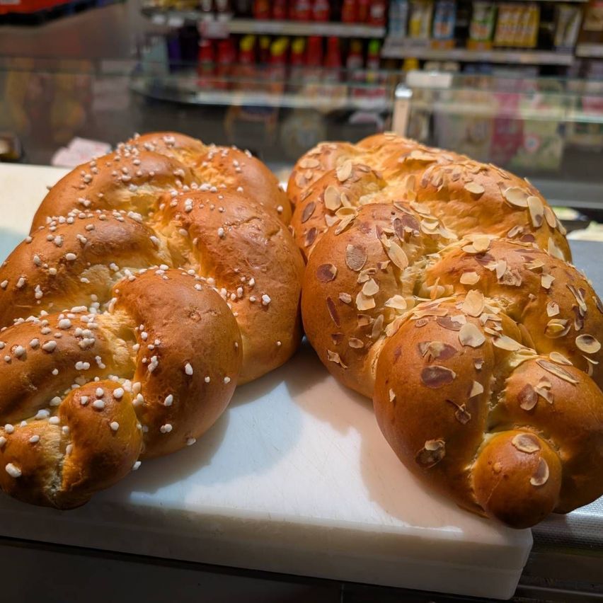 Two braided loaves of bread, one with white sesame seeds and the other with almonds, are on a white cutting board in front of a glass display case.
