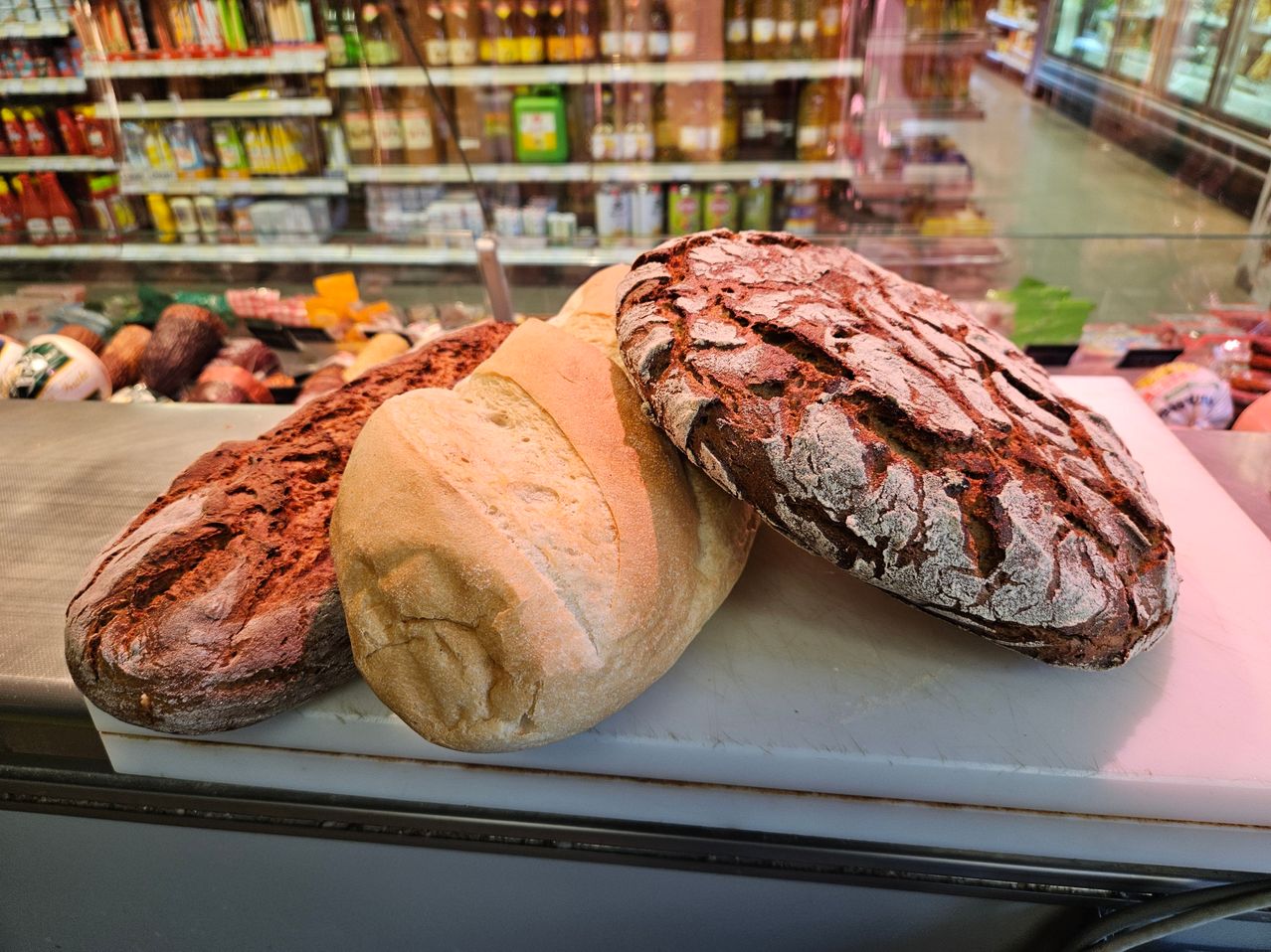 Three loaves of bread, including a light brown and two darker ones, are displayed on a counter in a shop with shelves of products in the background.