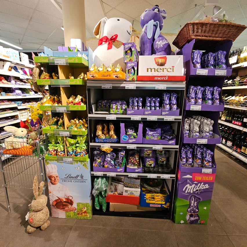 A supermarket aisle with shelves filled with various chocolate products, including Easter eggs and Lindt chocolates. Displayed prominently are purple and green boxes, and plush toys.