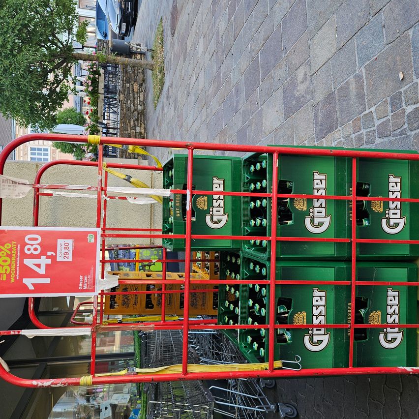 A red cart filled with green Günther beer crates, prominently displaying a red sign with a discount offer of 50% off, positioned against a brick wall with a tree in the background.