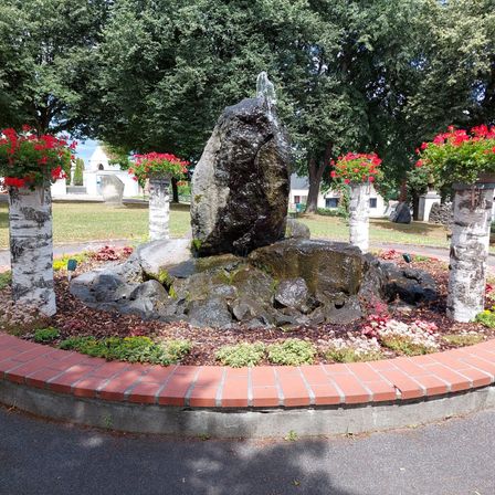 Ein Garten mit einem Brunnen aus Steinen und roten Blumen, umgeben von roten Ziegelkanten und Birken.
