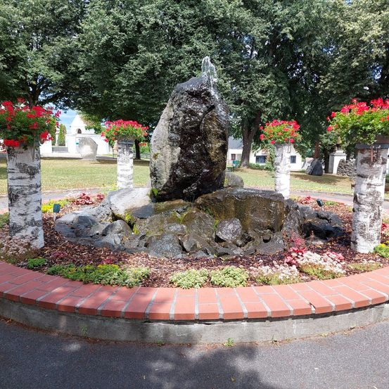 Ein Garten mit einem Brunnen aus Steinen und roten Blumen, umgeben von roten Ziegelkanten und Birken.