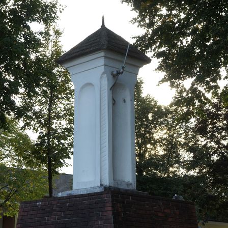 Ein weißer Turm mit Dach, auf einem Ziegelpodest montiert. Der Turm hat eine Metallleitung. Das Gebiet ist von Bäumen umgeben.