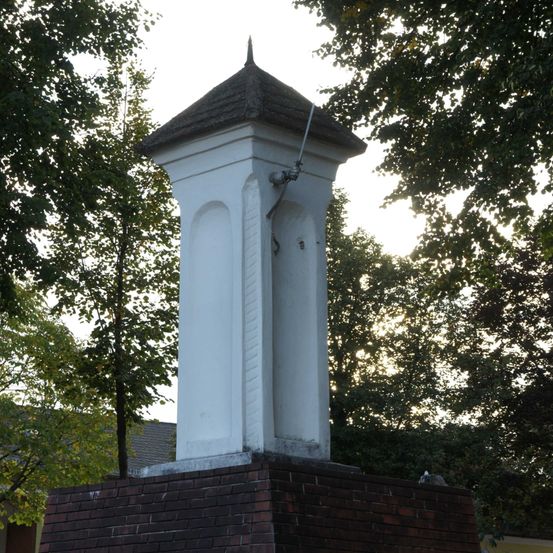 Ein weißer Turm mit Dach, auf einem Ziegelpodest montiert. Der Turm hat eine Metallleitung. Das Gebiet ist von Bäumen umgeben.
