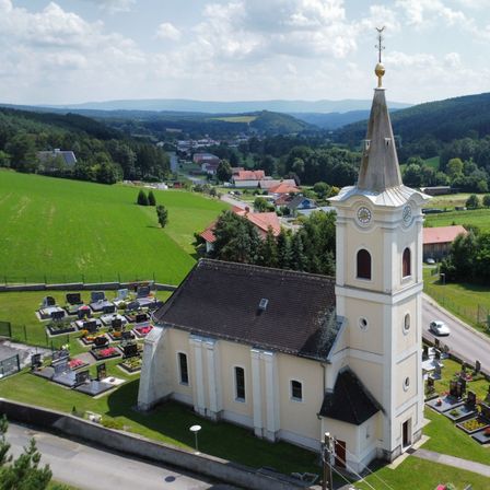 Luftaufnahme einer weißen Kirche mit Turm und Uhr, eingebettet in eine ländliche Landschaft mit grünen Feldern, einem Friedhof und einer Straße. Der Himmel ist teilweise bewölkt.