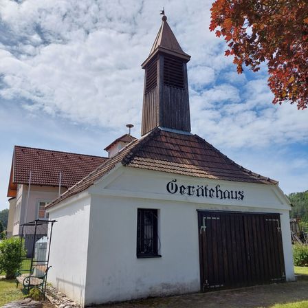 Ein weißes Gebäude mit braunem Dach und Turm, beschriftet mit "Gerätehaus", mit geschlossener Holztür, einem Fenster und einer Bank unter einem Baum davor.