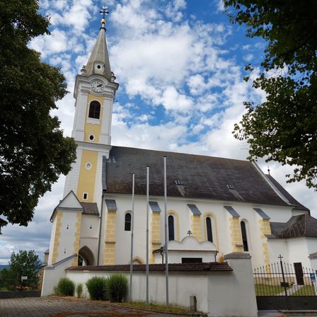 Eine weiße und gelbe Kirche mit Glockenturm und Turmspitze an einem sonnigen Tag mit blauem Himmel und weißen Wolken.