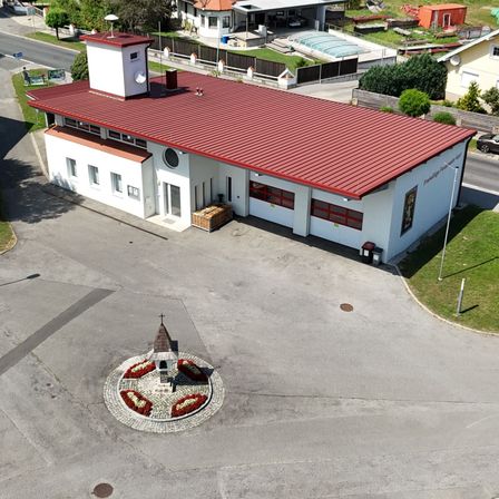 Luftaufnahme eines weißen Gebäudes mit rotem Dach und Schornstein, in einer kleinen Stadt gelegen. Es gibt Häuser, einen Swimmingpool und ein geparktes Auto in der Nähe.