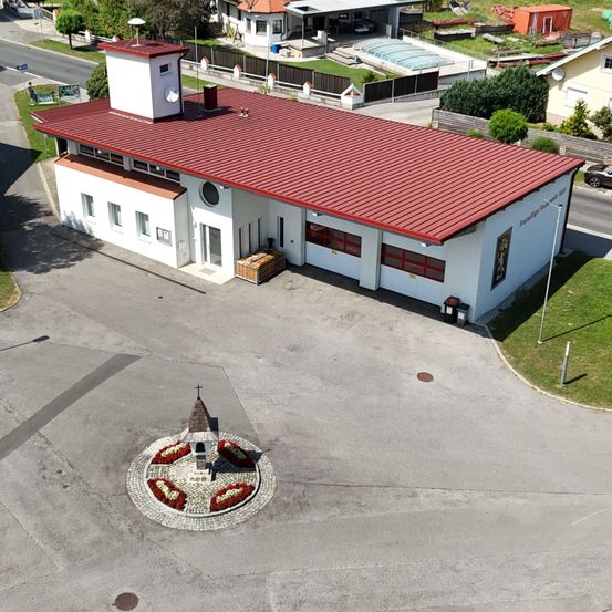 Luftaufnahme eines weißen Gebäudes mit rotem Dach und Schornstein, in einer kleinen Stadt gelegen. Es gibt Häuser, einen Swimmingpool und ein geparktes Auto in der Nähe.