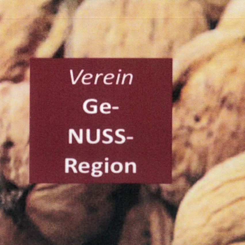 Ein Nahaufnahme eines Haufens Nüsse mit einem roten Schild, das 'Verein Ge- NUSS- Region' steht. Die Nüsse scheinen Walnüsse zu sein, und der Hintergrund ist leicht verschwommen.