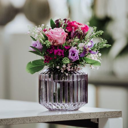 Eine Glasvase mit einer Blumenarrangement aus rosa, lila und weißen Blumen auf einem Betontisch.