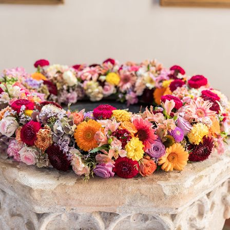 Ein farbenfroher Blumenkranz aus verschiedenen Blumen auf einem Steinsockel, vor einer weißen Wand.