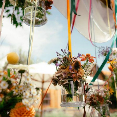 Eine Vielzahl von Blumen in Glasvasen hängen von Bändern unter einem klaren Himmel, mit einem Holzgebäude und einem weißen Schirm im Hintergrund.