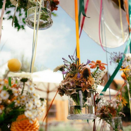 Eine Vielzahl von Blumen in Glasvasen hängen von Bändern unter einem klaren Himmel, mit einem Holzgebäude und einem weißen Schirm im Hintergrund.