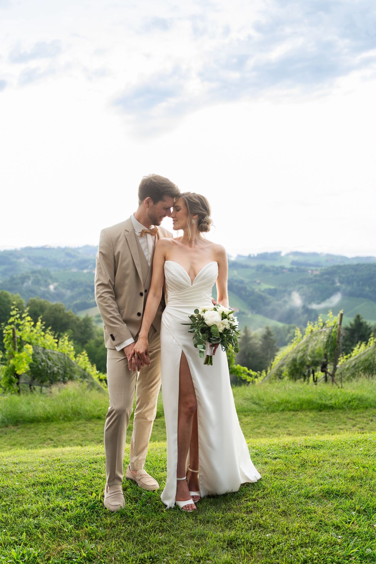 A newlywed couple stands on a grassy hill, the bride in a strapless white gown and the groom in a beige suit, both gazing into each other's eyes.