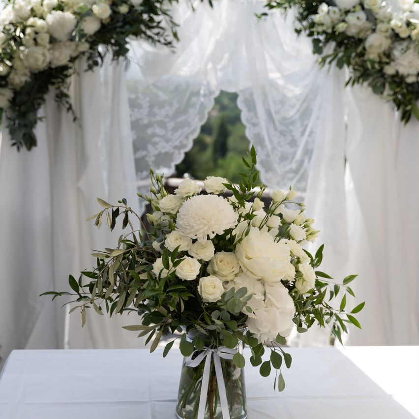 A white floral arrangement in a glass vase sits on a table, surrounded by white drapes and greenery.