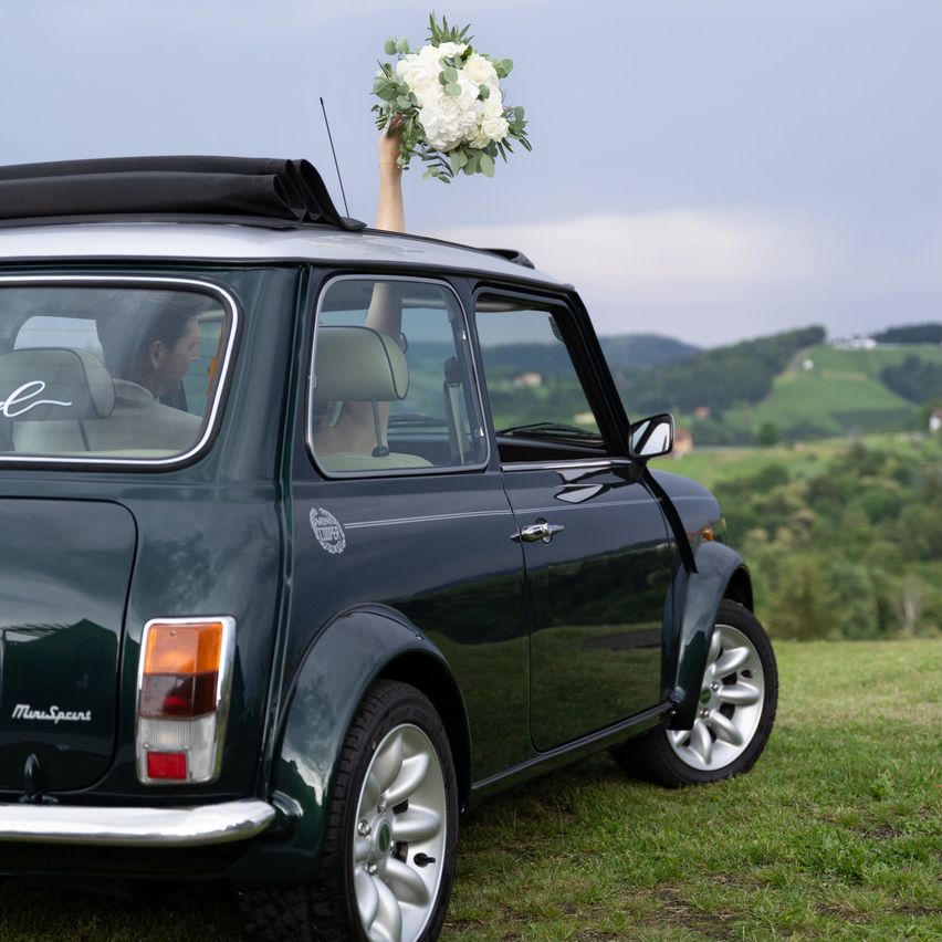 A bride holds a bouquet of flowers above the roof of a green Mini Cooper, parked on a grassy hill with a scenic view of mountains and greenery in the background.