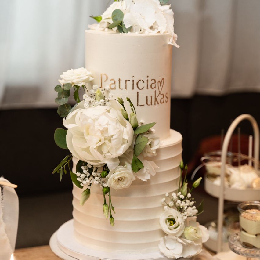 A three-tiered wedding cake with white flowers and green leaves, adorned with the names Patricia and Lukas in gold.