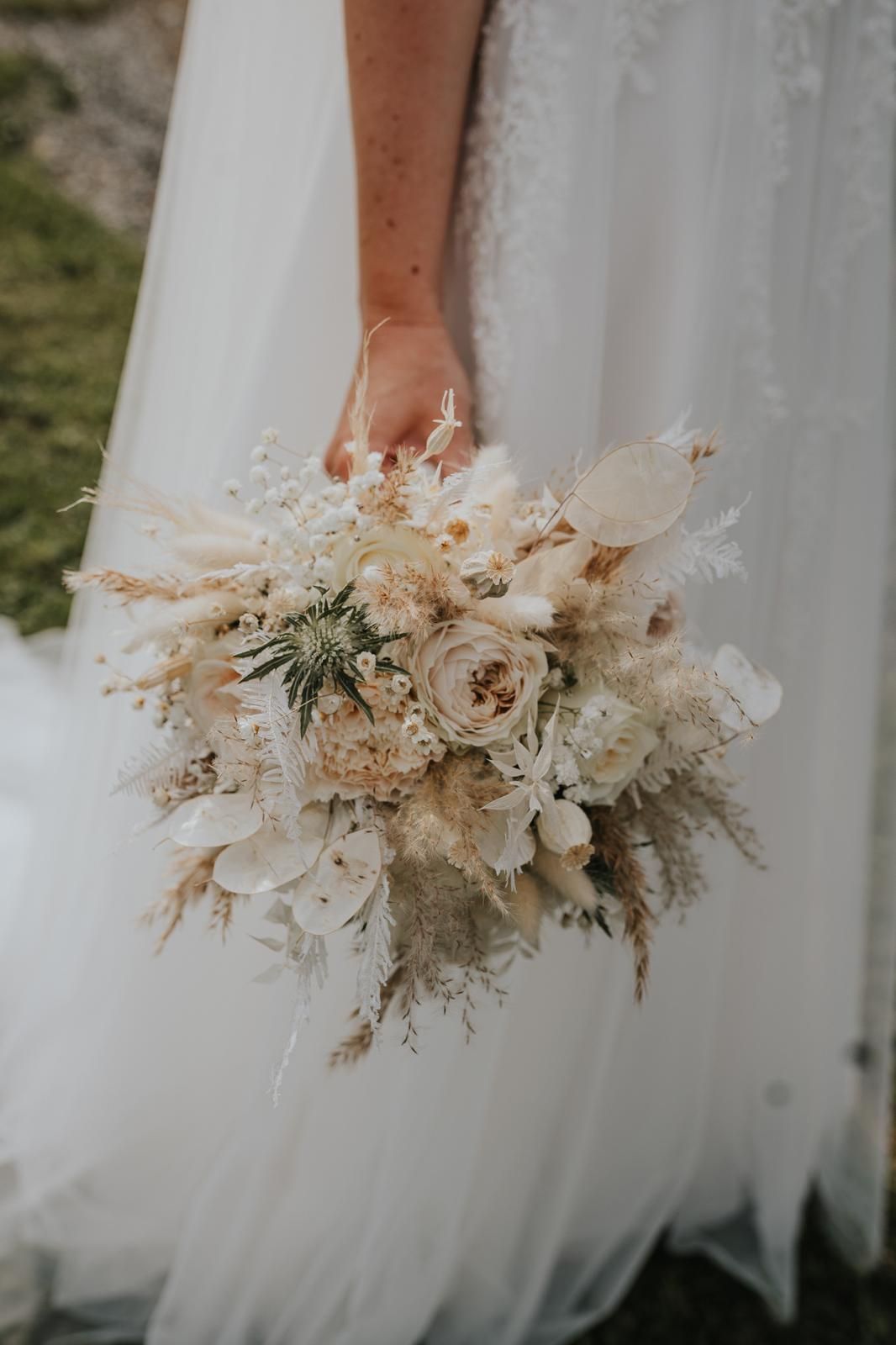 A bride in a white gown holds a wedding bouquet with white and cream flowers, dried leaves, and greenery.