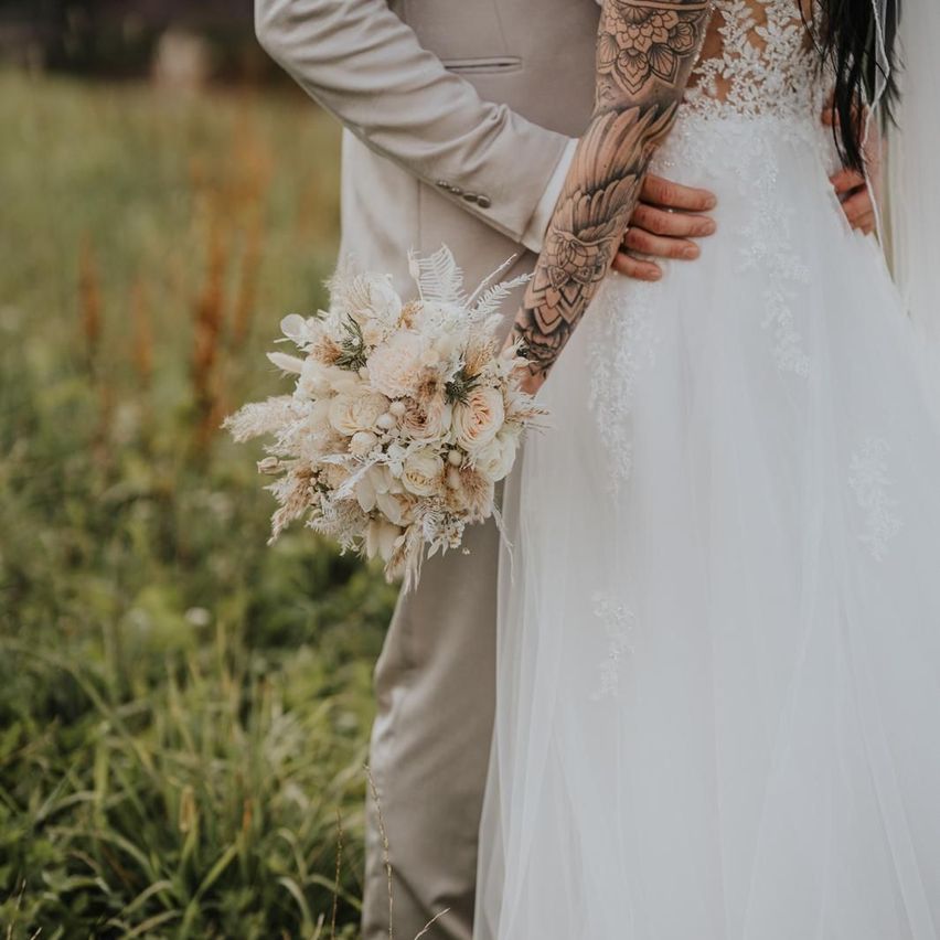 A bride in a white wedding dress holds a bouquet of white flowers while standing with her groom, who has tattoos on his arm.